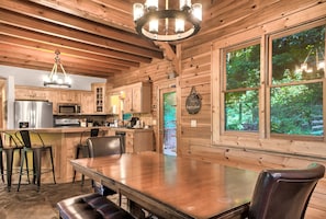 A wooden cabin interior with a kitchen, dining area, and large windows overlooking greenery.