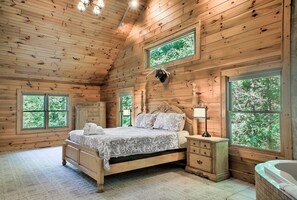 Ein Schlafzimmer in einer Holzhütte mit einem Bett, Nachttischen, einer Kommode und einem großen Fenster mit Blick auf Bäume.