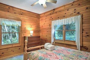 A wooden cabin room with a bed, a nightstand, a lamp, and two windows with curtains.
