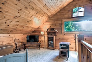 A cozy attic room with wooden walls, a television, a rocking chair, a bookshelf, and a window with a view of trees.