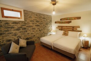 A bedroom with two beds, a wooden headboard, a stone wall, a framed picture, a sofa, and a nightstand with a lamp.