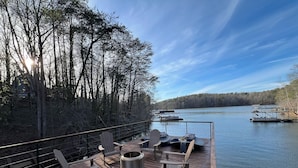 Eine Holzterrasse mit Adirondack-Stühlen und einem Feuerkorb mit Blick auf einen ruhigen See und Boote.