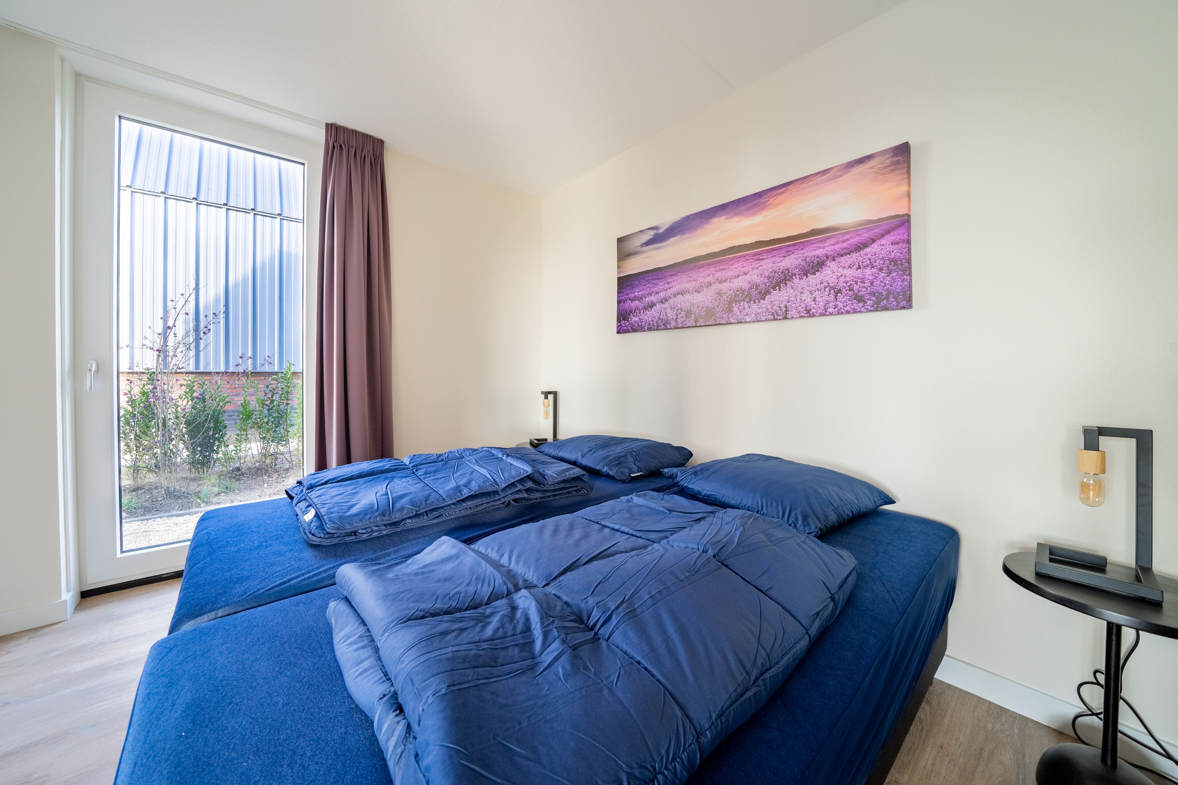 A bedroom with a large bed, a window with curtains, a painting on the wall, and a small table with a lamp.