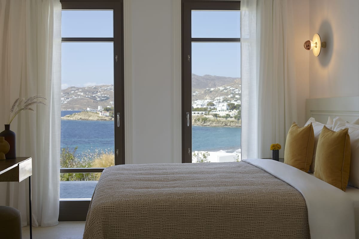 A hotel room with a bed, a nightstand, and a view of the sea and buildings.