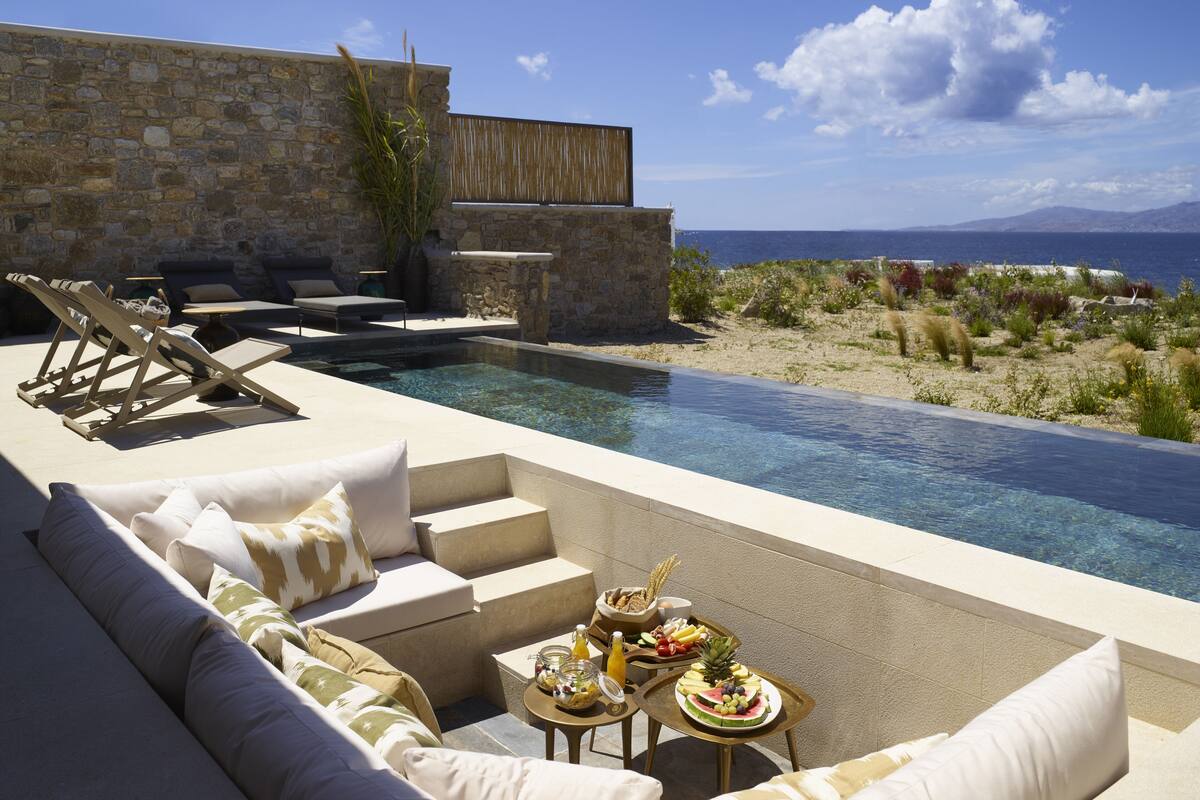 An infinity pool with a stone wall and lounge chairs, a dining table with fruit and drinks, and a view of the sea.