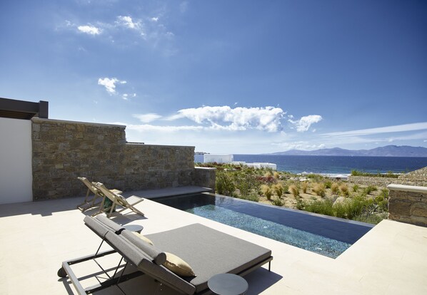 A modern outdoor pool area with lounge chairs and a stone wall, overlooking a scenic coastline. - Bonzoe Suites & Villas (Mykonos)
