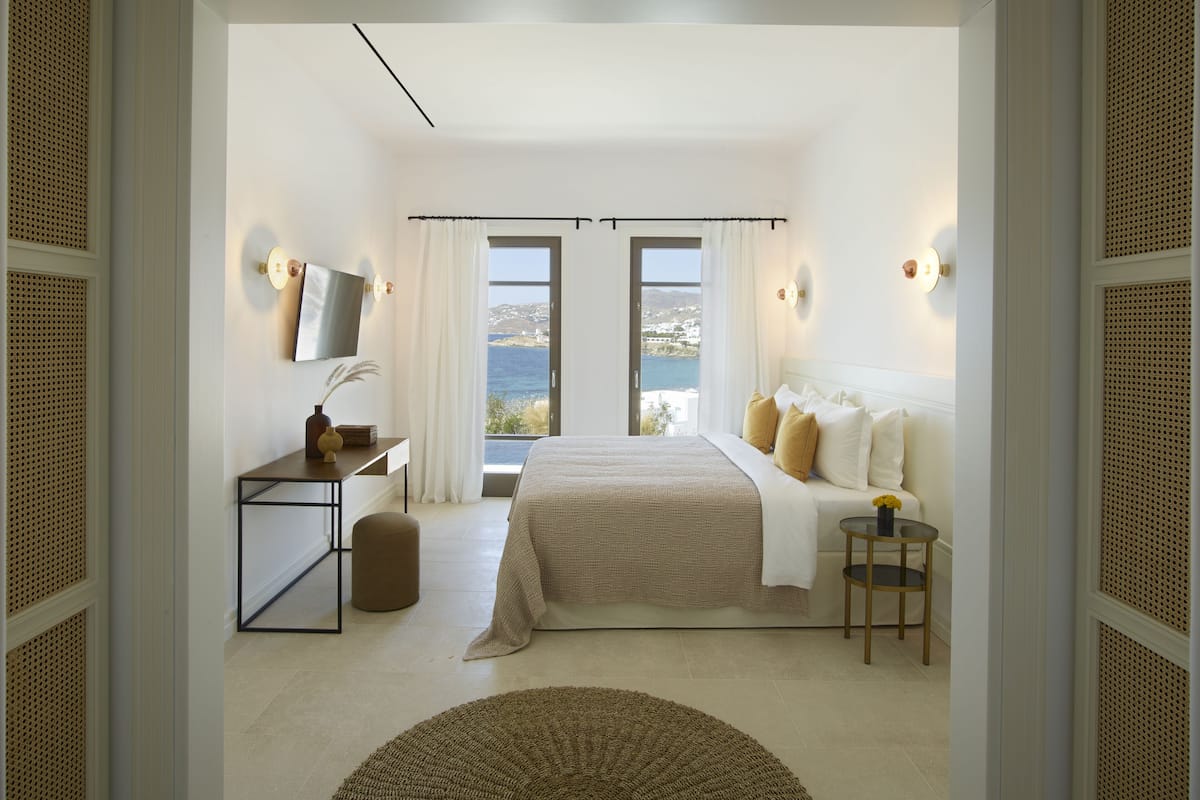 A bedroom with a bed, a desk, a chair, and a view of the sea.