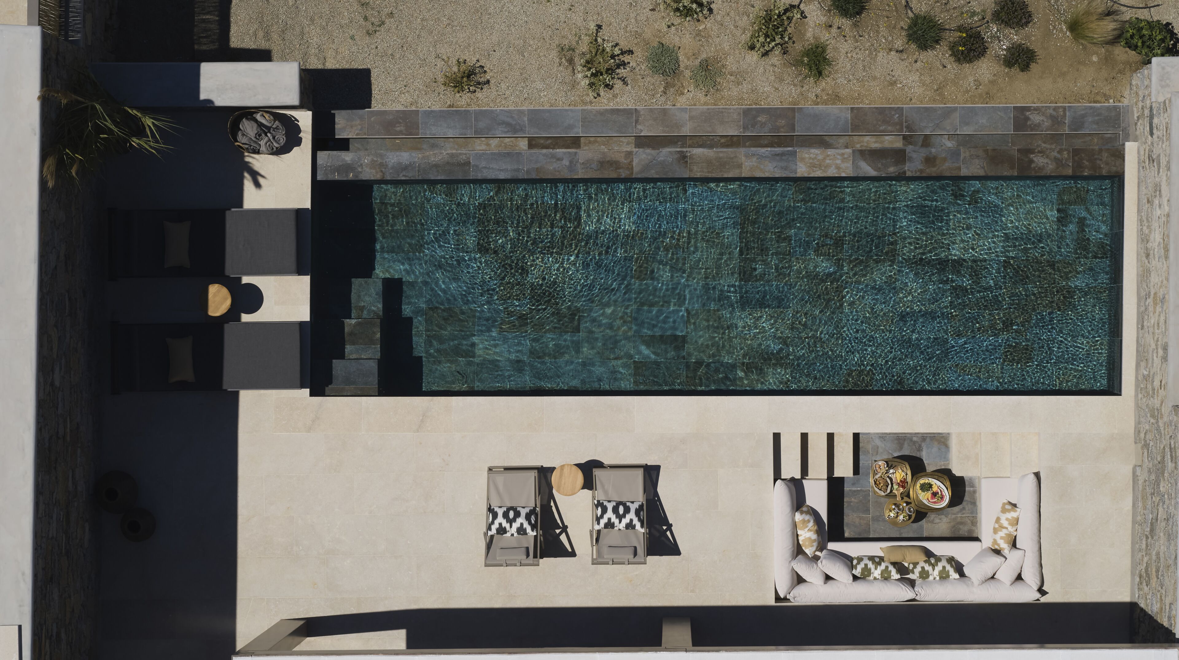 An aerial view of a modern outdoor swimming pool area with a sun lounger, a small table with a vase, and a seating area with cushions.