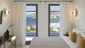Et soveværelse med en stor seng, to døre der fører til en balkon med udsigt over havet og bygninger.