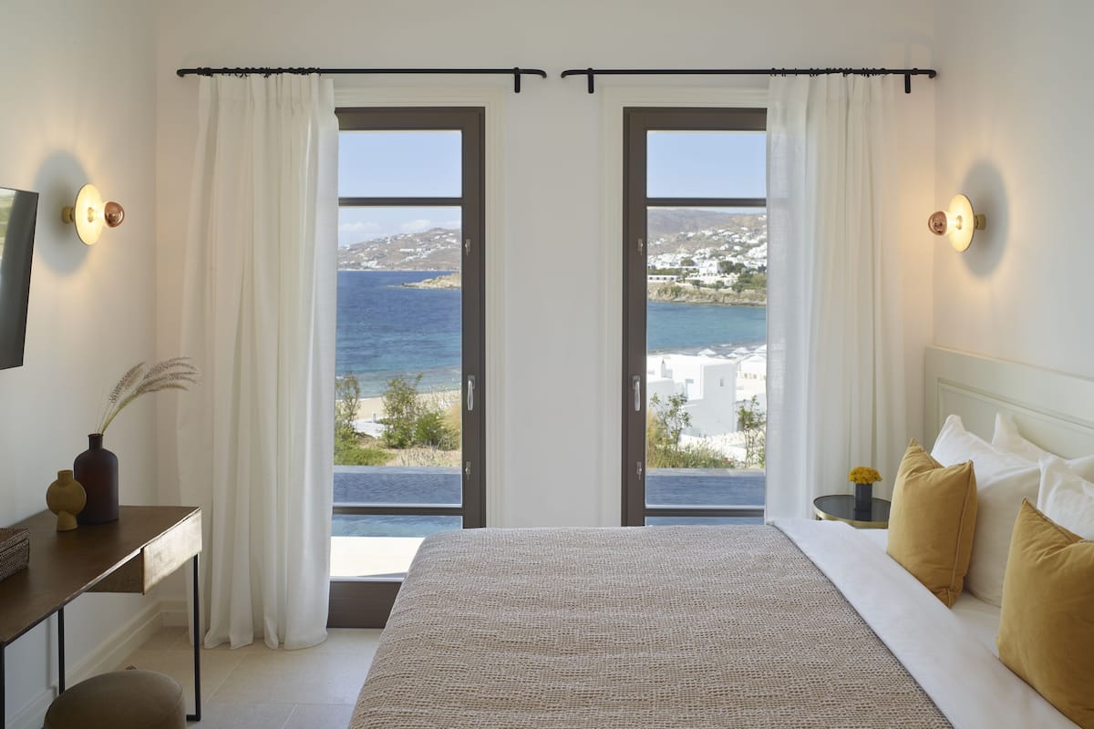 A bedroom with a large bed, two doors leading to a balcony with a view of the sea and buildings.
