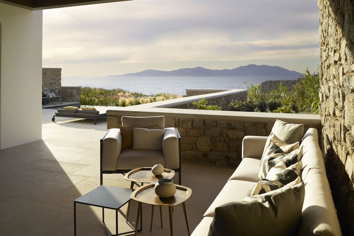 A modern outdoor seating area with a stone wall, a sofa, and two chairs, overlooking a scenic view of the sea and mountains.