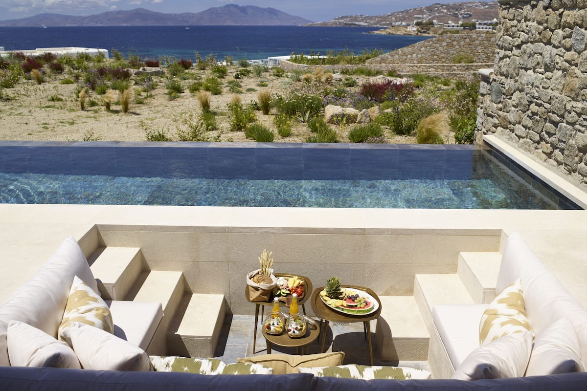 A poolside area with a view of the sea and mountains, a stone wall, and a table with food and drinks.