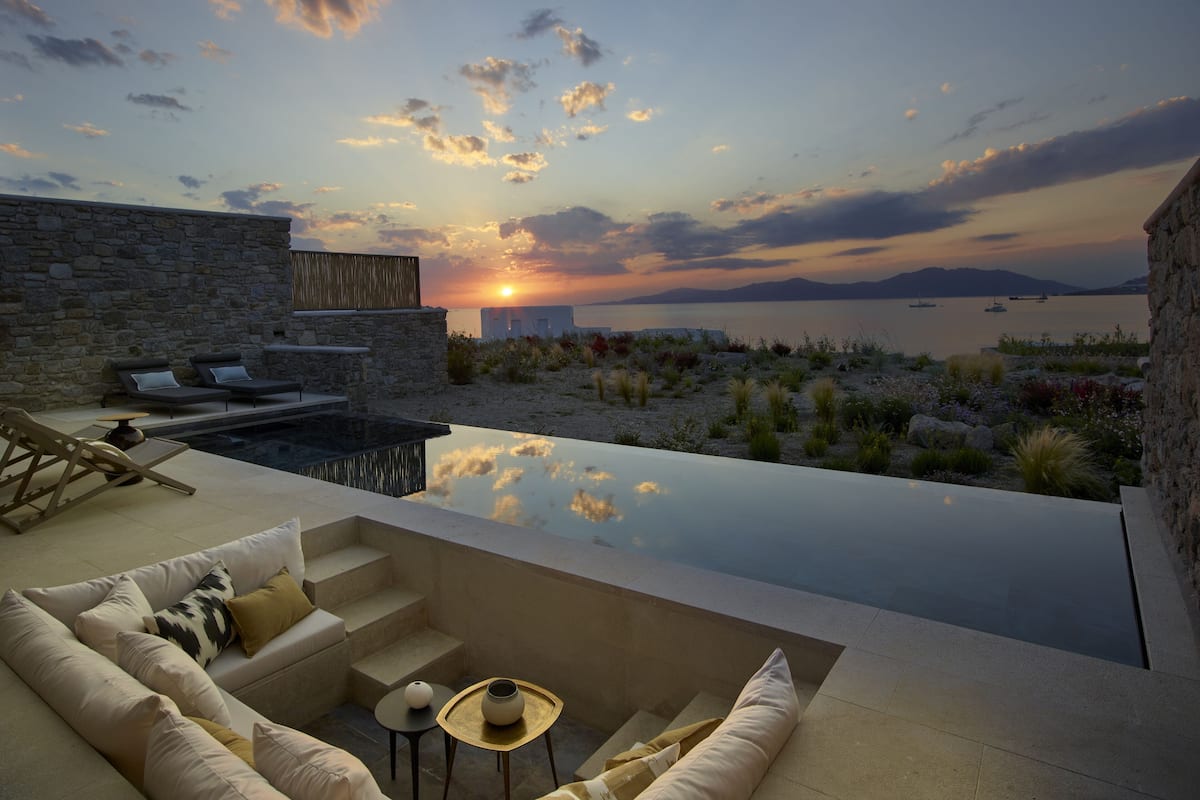A modern outdoor lounge area with a stone wall, a pool, and a view of the sunset over the sea.