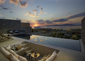 A modern outdoor lounge area with a stone wall, a pool, and a view of the sunset over the sea. - Bonzoe Suites & Villas (Mykonos)