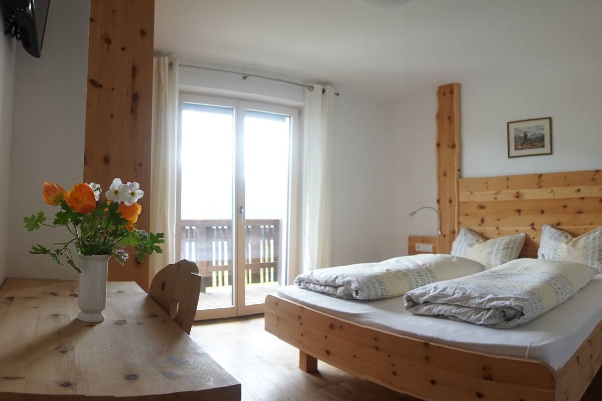 A bedroom with a wooden bed, a wooden nightstand, a bedside table with a vase of flowers, and a sliding glass door to the balcony.