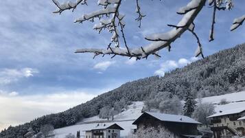 Een besneeuwd berglandschap met gebouwen, waaronder een berghut en een ruimere woning met balkon, omringd door bomen en een heldere lucht.
