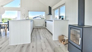 Moderni keittiö, jossa on valkoiset kaapit, keskellä oleva keittiösaareke, liesi ja suuret ikkunat, joista avautuu näkymä ulos.