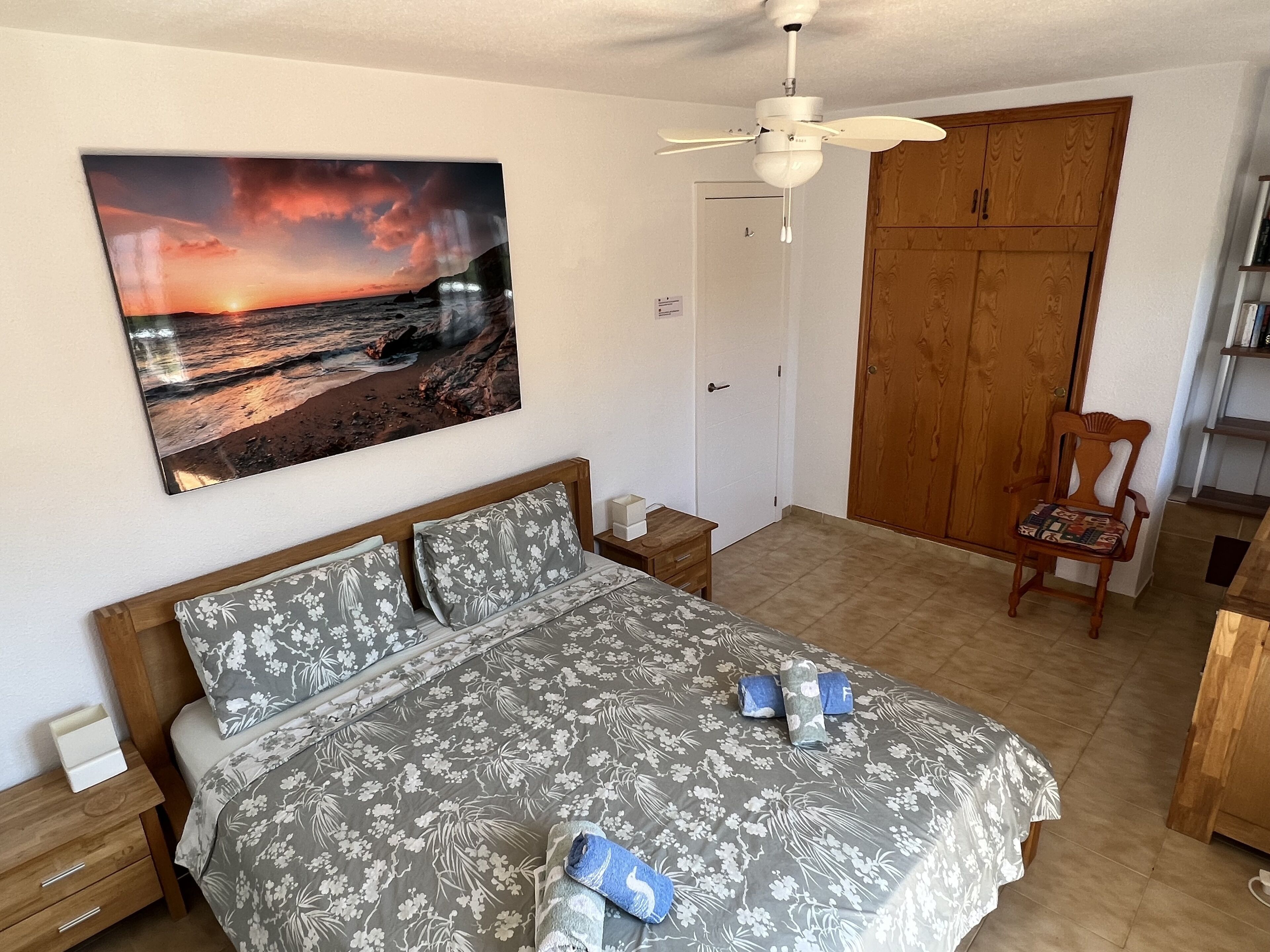 A bedroom with a bed, a wooden wardrobe, a chair, a ceiling fan, and a painting of a sunset over the ocean.