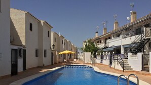 A swimming pool with a tiled deck, surrounded by white buildings with balconies and a yellow umbrella.