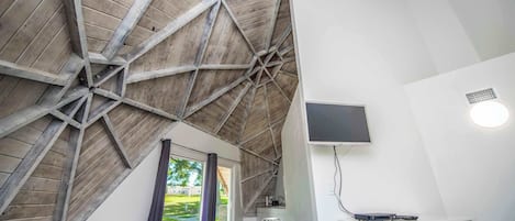 Ein Zimmer mit schrägen Wänden, einem an der Wand befestigten Flachbildfernseher, einem Schreibtisch mit Stuhl und einem Fenster mit Blick auf Grünfläche.