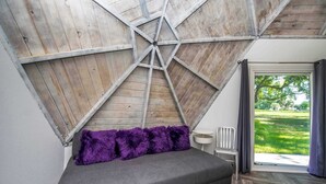 Ein Zimmer mit schräg verlaufender Decke, ein graues Sofa mit violetten Kissen, ein weißer Sessel und der Blick auf eine grüne Landschaft durch ein großes Fenster.