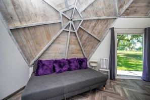 A room with a sloped ceiling, a grey sofa with purple cushions, a white chair, and a view of a green landscape through a large window.