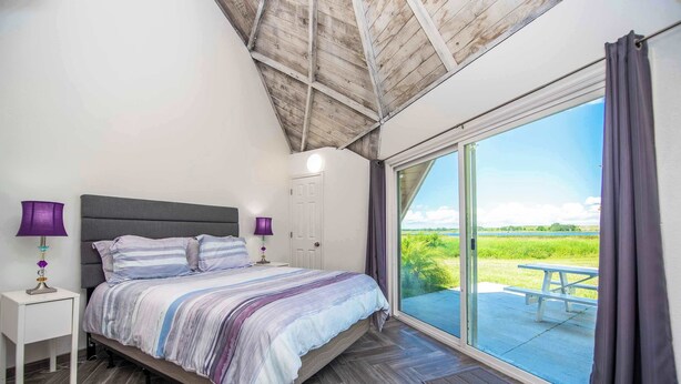 Ein Schlafzimmer mit einem großen Bett, zwei Nachttischen, einem Fenster mit Vorhängen und Blick auf eine Rasenfläche.