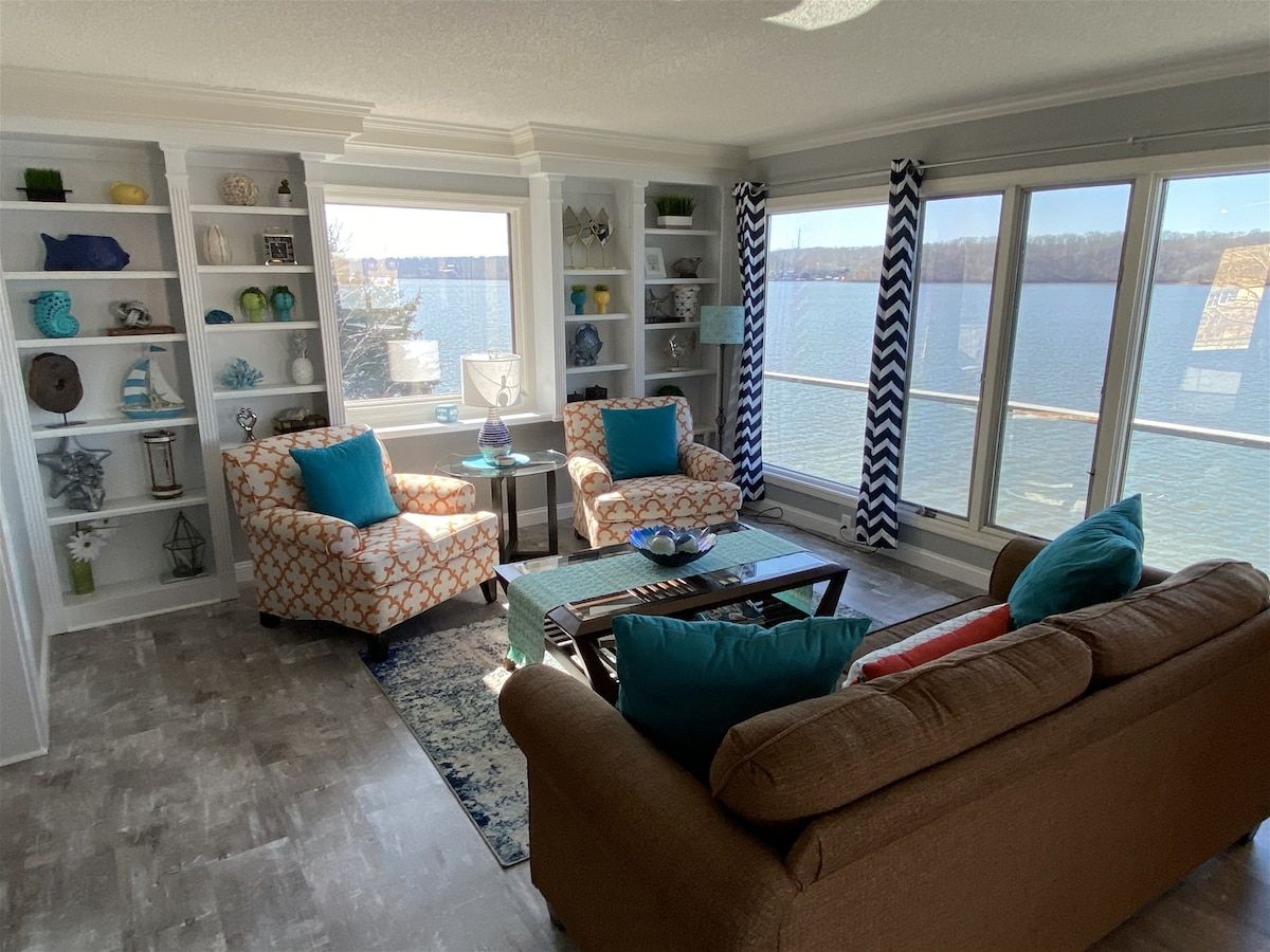 A living room with a view of a body of water, furnished with a brown sofa, patterned armchairs, and a coffee table.