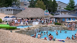 Un espace piscine doté de plusieurs éléments aquatiques, notamment une piscine centrale et un espace avec un jacuzzi, le tout entouré d’une plage de galets.