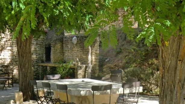 An outdoor dining area with a stone table and metal chairs, surrounded by trees and a stone wall. -  Air Conditioning House with character located  in Luberon (Ménerbes)