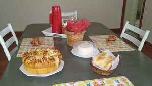 A dining table with a red coffee maker, a basket of flowers, and a cake on a floral tablecloth. - Pousada do joao (Juiz de Fora)