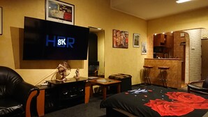 Ett hotellrum med en stor platt-TV, en soffa, ett litet bord, en fläkt, en säng med ett rött, blommigt sängöverkast och en liten köksdel.