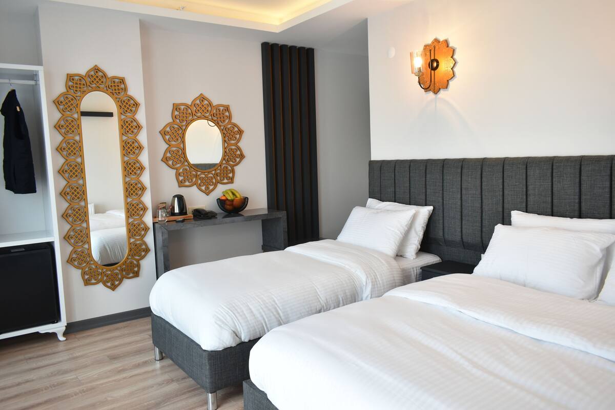 A hotel room with two beds, a large mirror, a wall-mounted light fixture, and a small table with a fruit bowl.