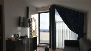 A modern living room with a dark sofa, a flat-screen TV mounted on the wall, and a view of the sea through a large window with dark curtains. - Avcilar Garden Hotel (Istanbul)
