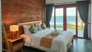 Une chambre avec un mur en briques, un lit en bois, une table de chevet, une lampe et une vue sur la mer par une porte vitrée.