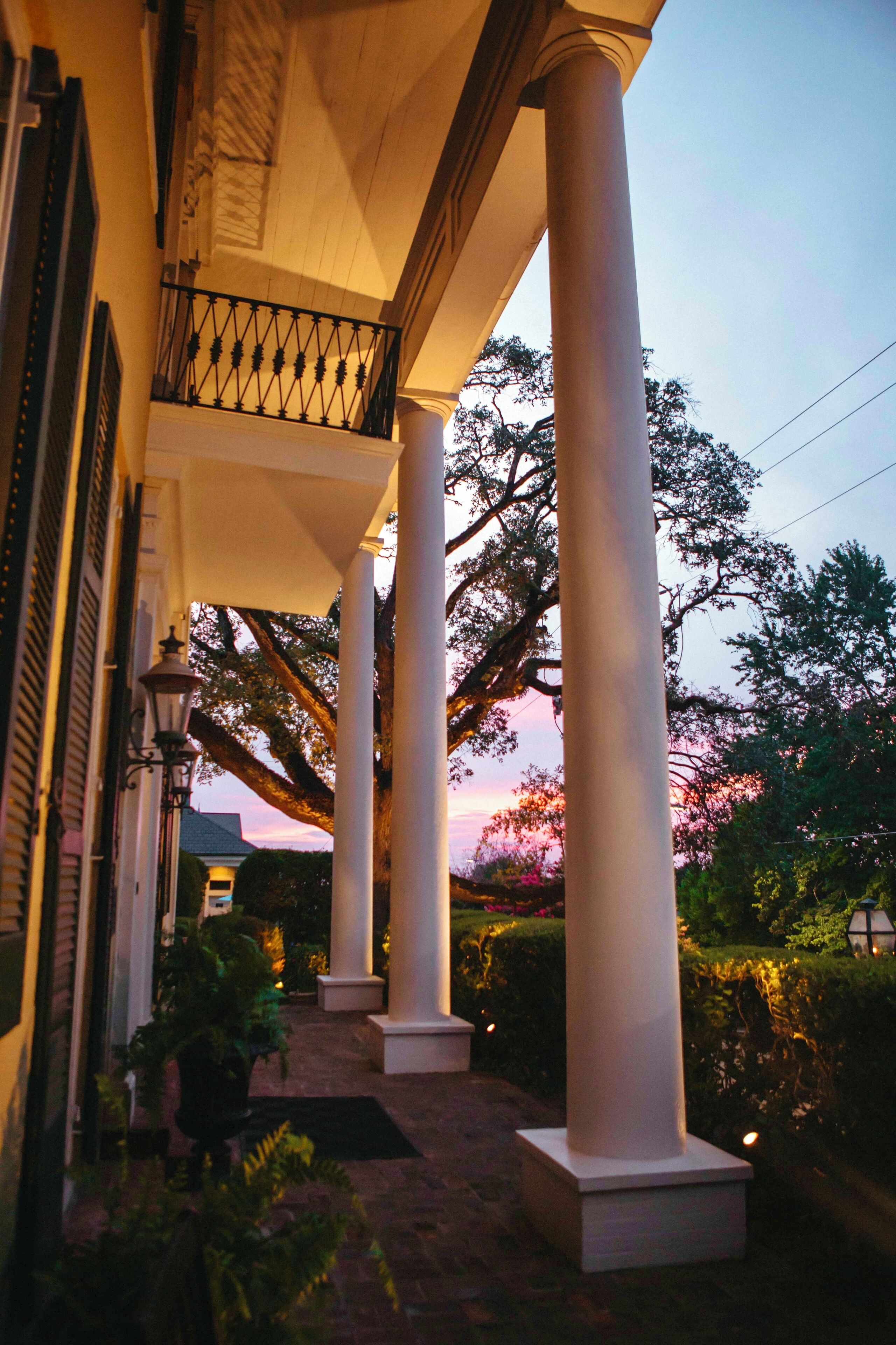 A covered outdoor area with columns and a railing, surrounded by greenery and a sunset in the background.