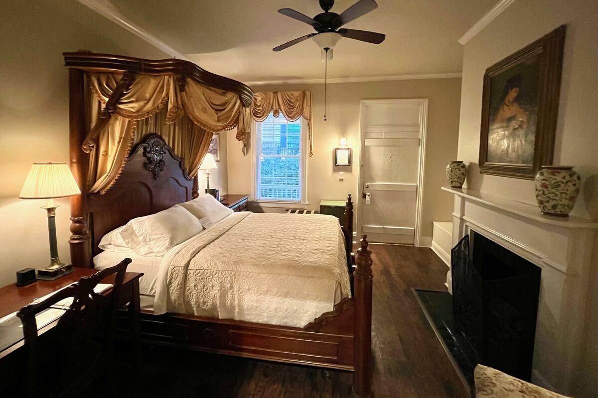 A bedroom with a four-poster bed, a ceiling fan, a fireplace, and a view of the city through the window.