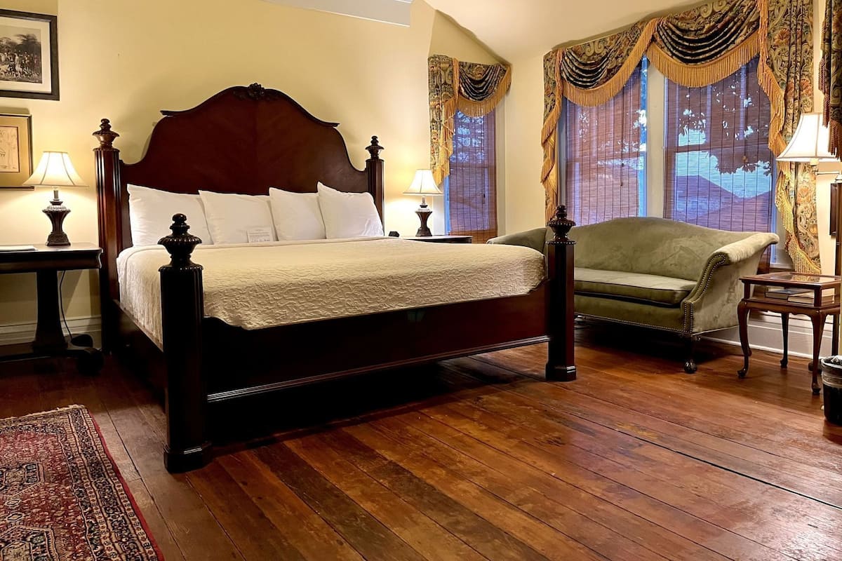 A traditional four-poster bed with a wooden headboard, a green armchair, and a wooden nightstand with a lamp.