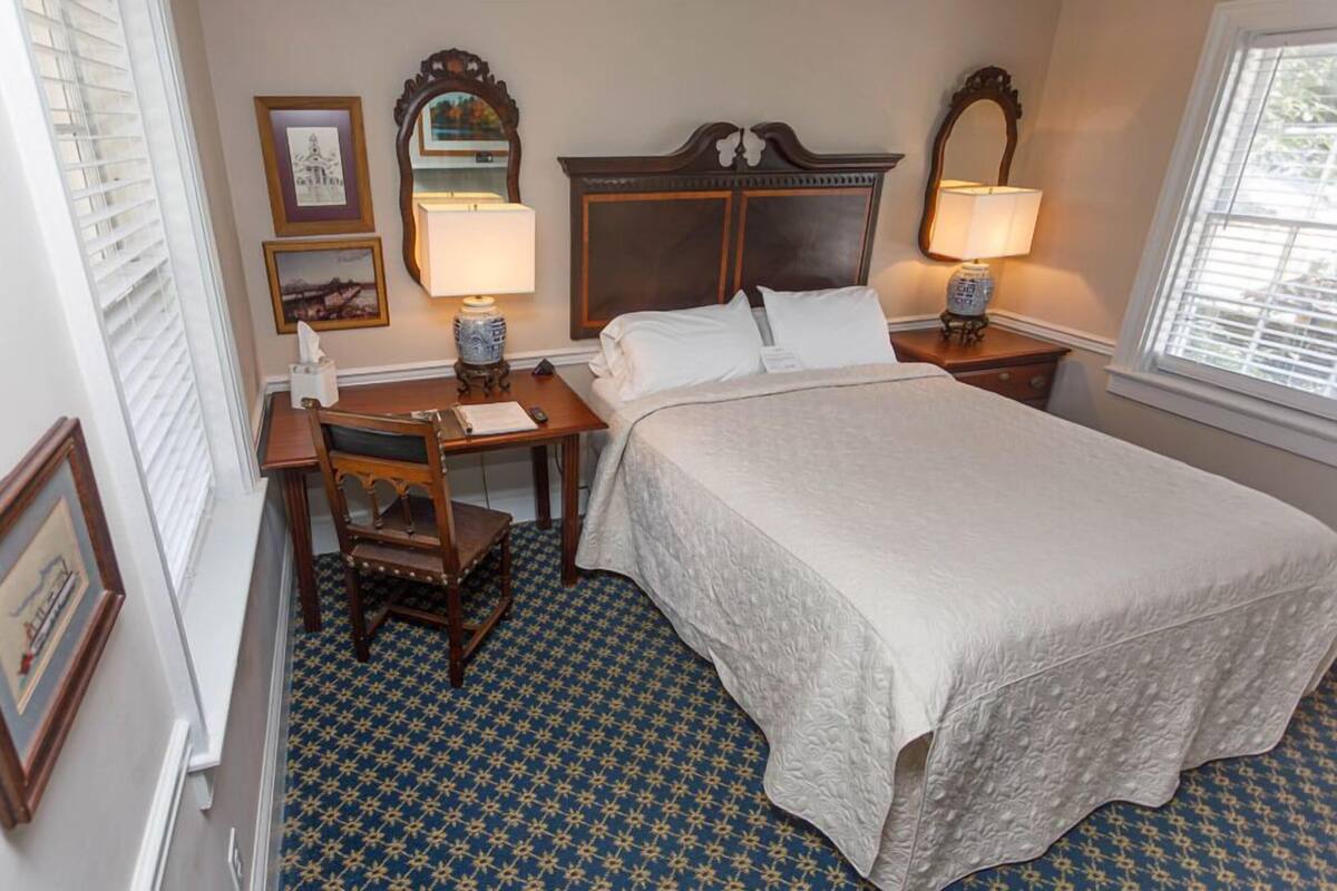 A hotel room with a large bed, two bedside tables with lamps, a wooden chair, a desk, and a patterned carpet.