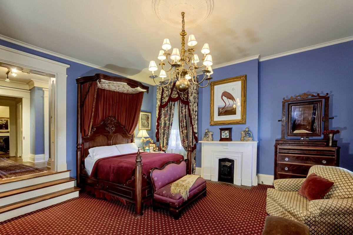 A bedroom with a four-poster bed, a chandelier, a fireplace, and a sofa.