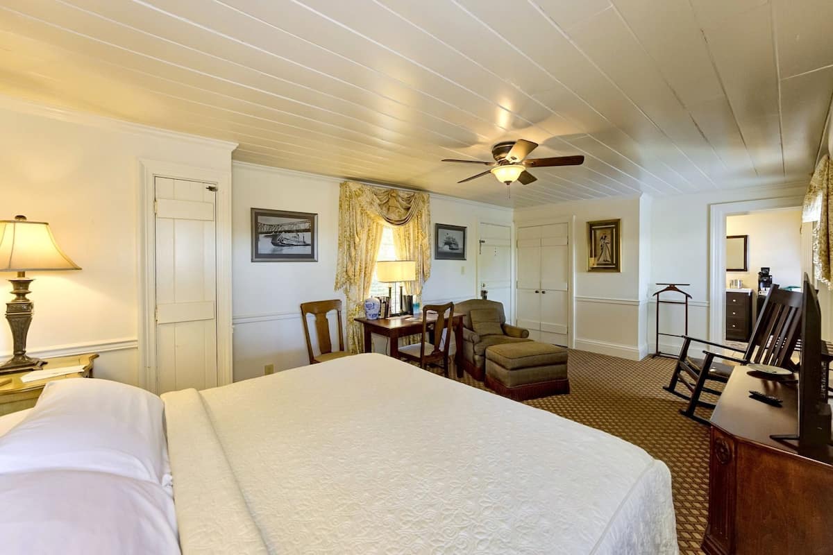 A hotel room with a bed, a desk, a chair, a desk lamp, a ceiling fan, and a wardrobe.