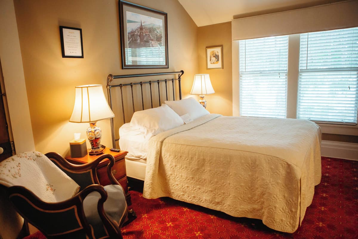 A hotel room with a bed, two bedside lamps, a chair, a desk with a lamp, and a window with blinds.