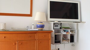 Huoneessa on puinen kaappi, seinälle asennettu televisio, lamppu sekä hylly, jossa on kirjoja ja elektronisia laitteita.