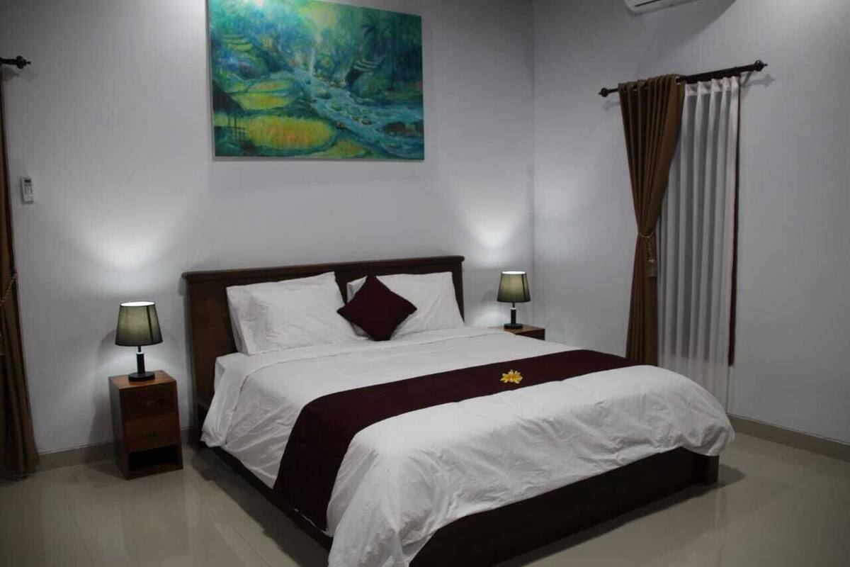 A neatly made bed with white bedding and a maroon throw, flanked by two bedside tables with lamps, in a room with a large abstract painting on the wall and a window with curtains.