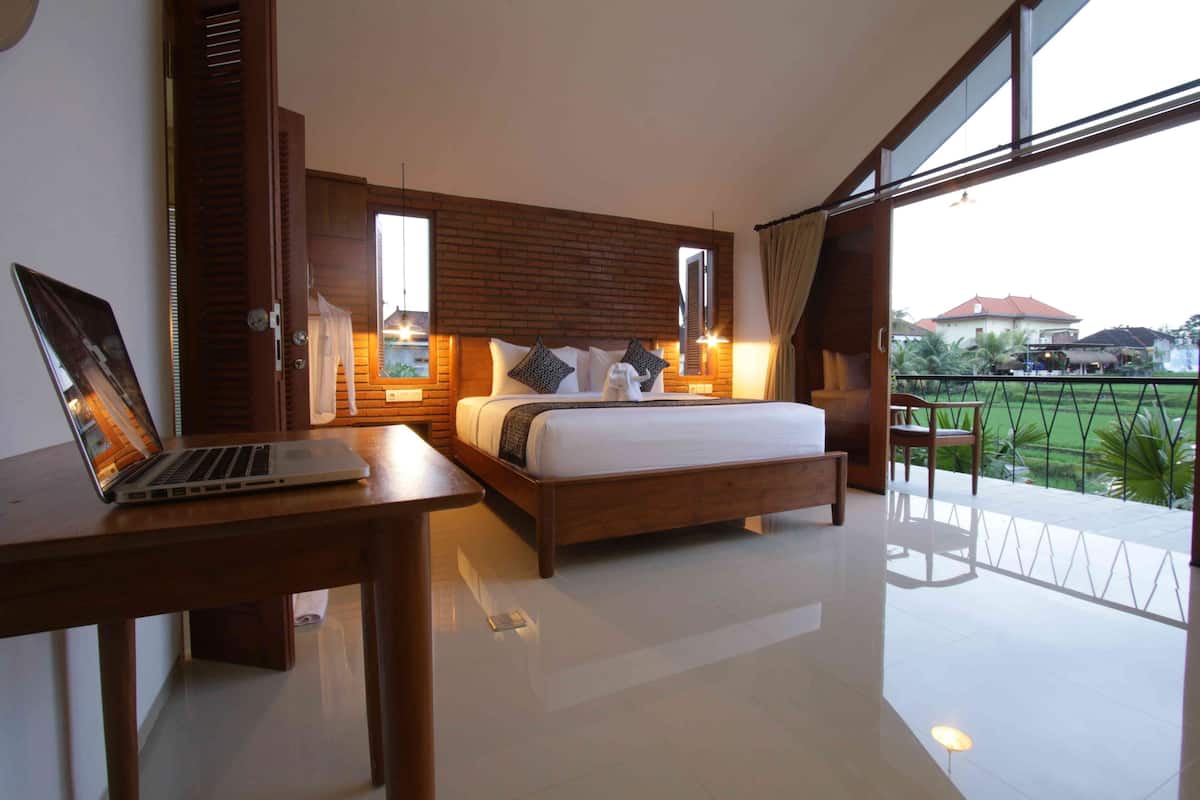 Quarto moderno com uma cama grande, um laptop numa mesa de madeira e uma varanda com vista para os arredores.