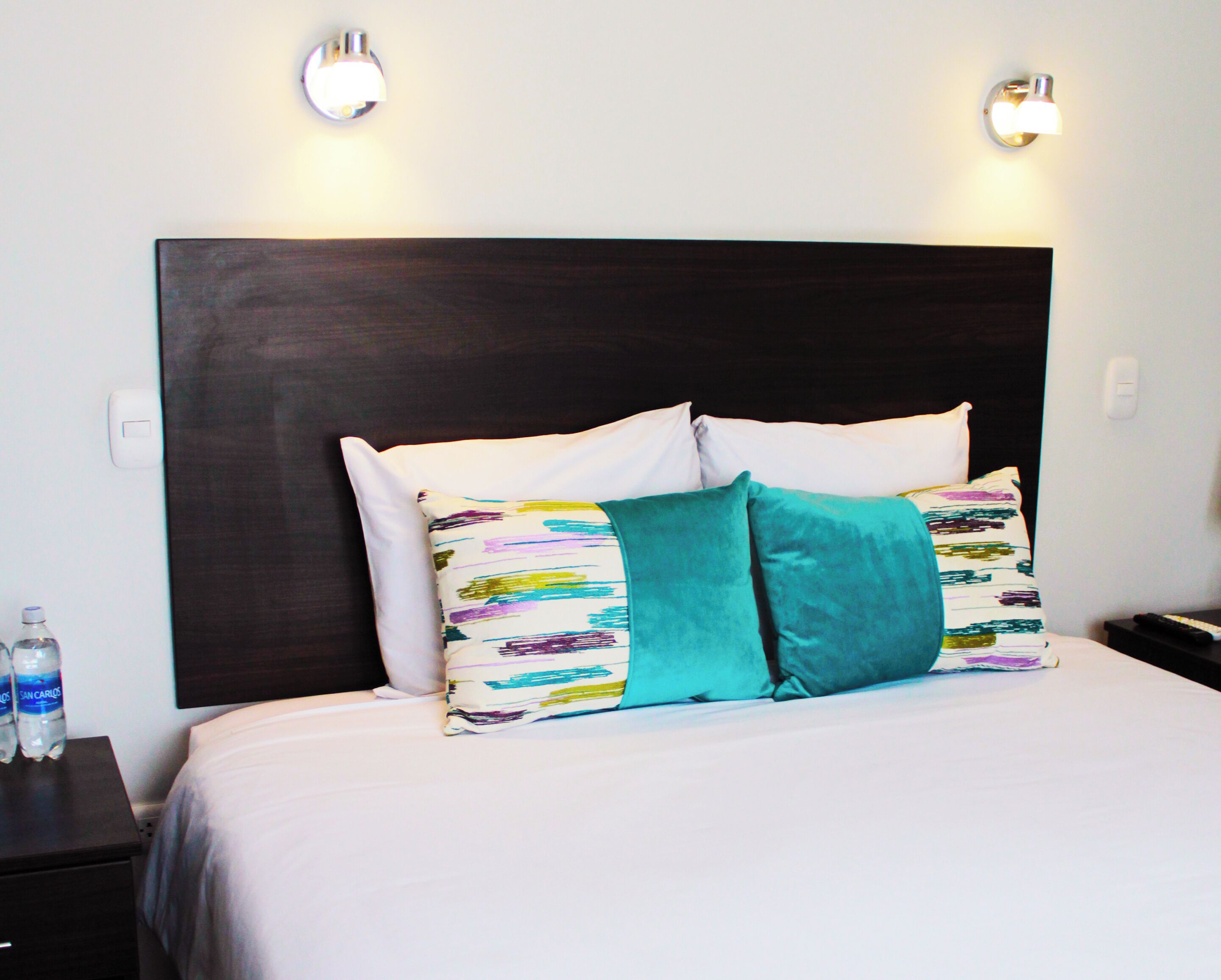 Una cama bien tendida con ropa de cama blanca y almohadas estampadas y coloridas, un cabecero oscuro y dos lámparas fijadas a la pared.