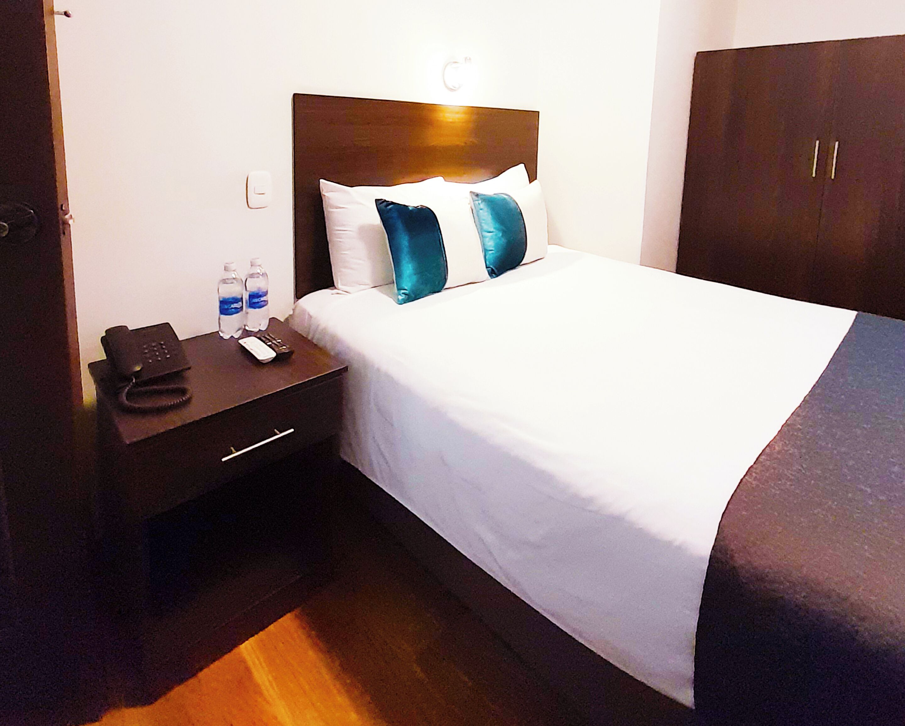 A hotel room with a bed, a nightstand with a phone and remote, a water bottle, and a wardrobe.