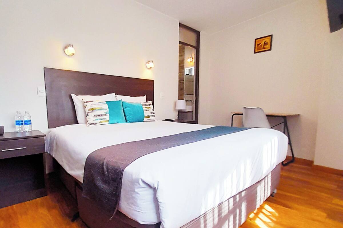 Habitación de hotel con una cama grande, un escritorio y una silla. Hay dos botellas de agua sobre la mesita de noche.