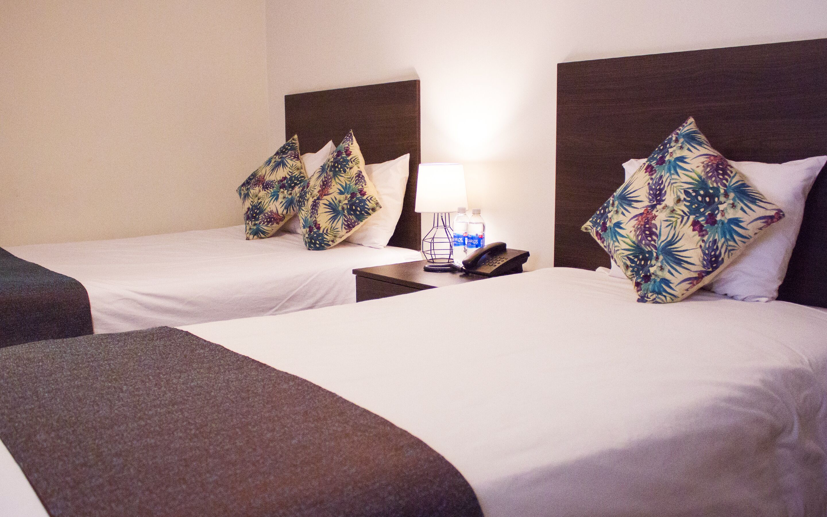 A hotel room with two beds, each with floral-patterned pillows, a bedside table with a lamp and a phone, and a water bottle.