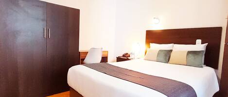 Habitación de hotel con una cama grande, un armario, un escritorio y una mesita de noche con lámpara.